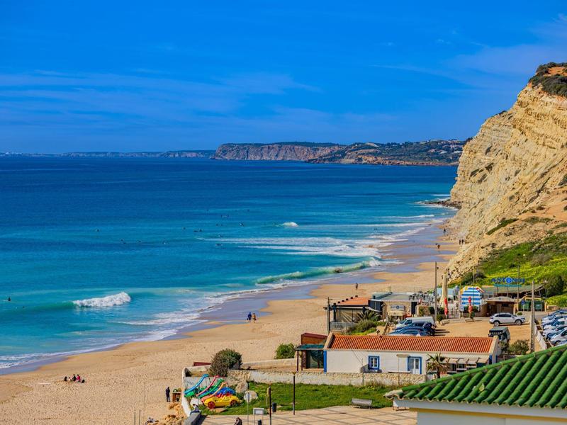 Strand mit goldgelbem Sand, blauen Wellen und hohen Klippen unter hellem blauem Himmel.