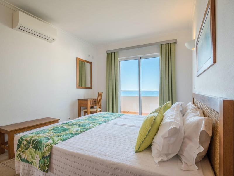 Hell eingerichtetes Schlafzimmer mit Doppelbett, grünem Vorhang und Meerblick durch die Tür.