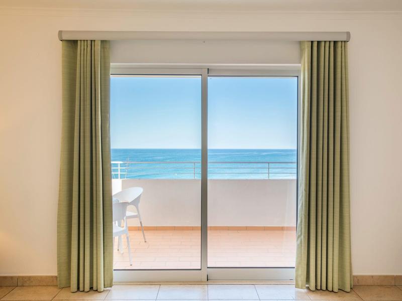 Großes Fenster mit grünen Vorhängen, Blick auf Terrasse und blaues Meer am sonnigen Tag.