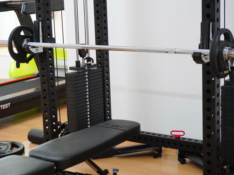 Schwarze Hantelbank und Langhantel mit Gewichten in einem hellen Fitnessraum mit Holzboden.