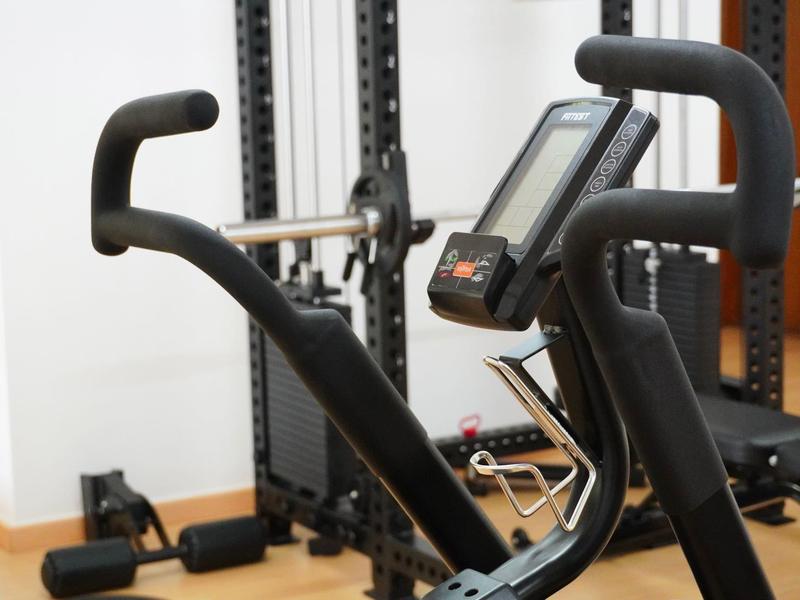 Schwarzes Fitnessgerät mit Griffen und digitalem Display in hellem Raum mit Holzfußboden.