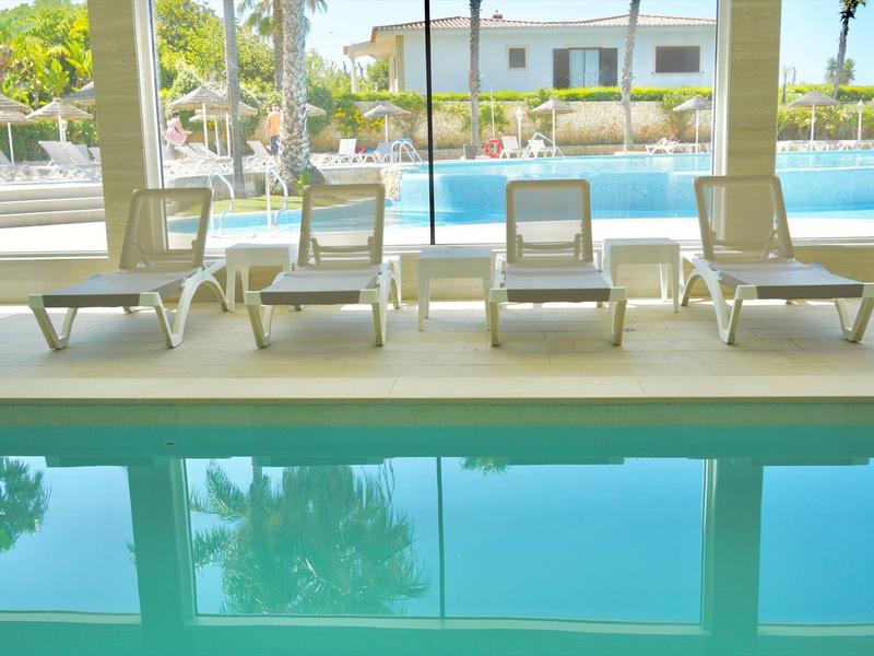 Innen- und Außenpool mit mehreren weißen Liegestühlen, Palmen und hellem Gebäude im Hintergrund.