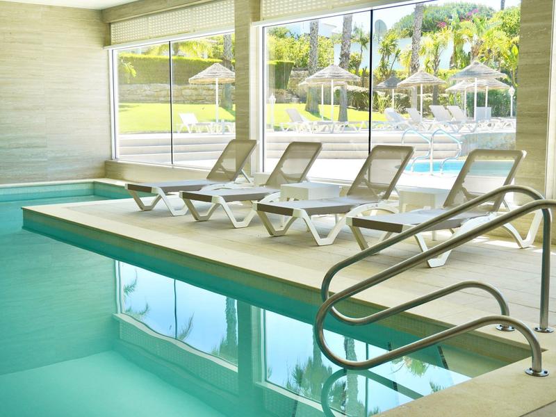 Hell beleuchteter Innenpool mit Liegestühlen neben großen Fensterfronten und Blick ins Freie.