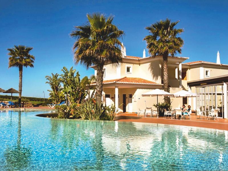 Großer Pool mit Palmen vor einem hellen Hotelgebäude an einem sonnigen Tag.