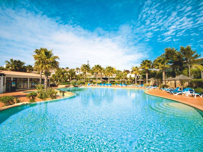 Großer Hotelpool mit Palmen und Sonnenliegen unter blauem Himmel.