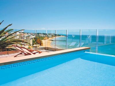 Blauer Pool mit Glasgeländer, Liegestühle und Palmen, Meer und Strand im Hintergrund bei klarem Himmel.
