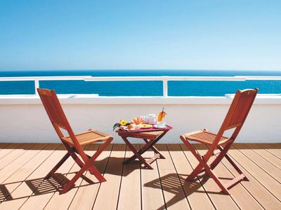 Zwei Holzkissenstühle mit kleinem Tisch auf heller Terrasse mit Meer und blauem Himmel im Hintergrund.