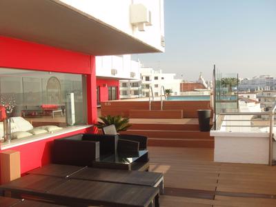 Moderne Dachterrasse mit Holzdeck, Glasgeländer, schwarzer Lounge-Möbel und roter Wand.