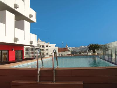 Moderner Pool auf Dachterrasse mit Glasgeländer, Sonnenliegen, hellem Gebäude und klarem blauem Himmel.