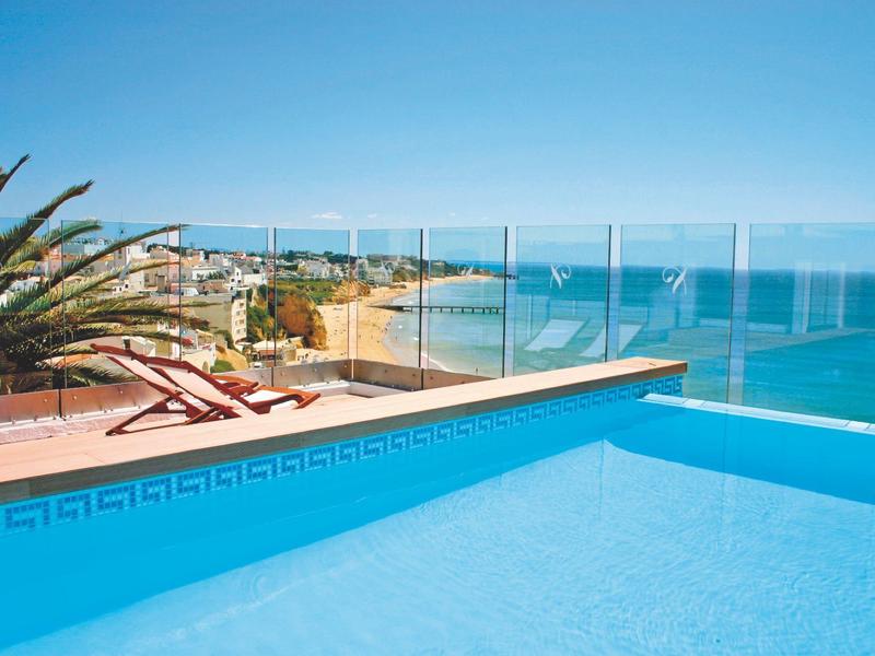 Blauer Pool mit Glasgeländer, Liegestühle und Palmen, Meer und Strand im Hintergrund bei klarem Himmel.