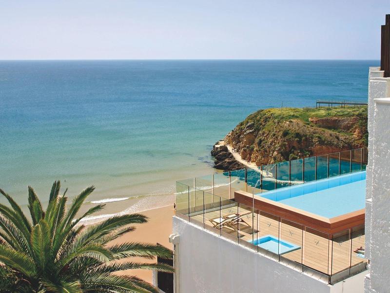Schwimmbad an der Küste mit Blick auf den Strand, das blaue Meer und einen bewachsenen Felsen.