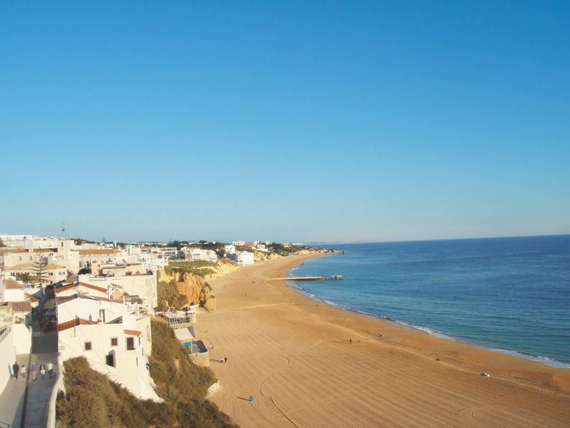 Strand mit goldenem Sand, blauem Himmel und Meer, daneben weiße Häuser am Küstenrand.