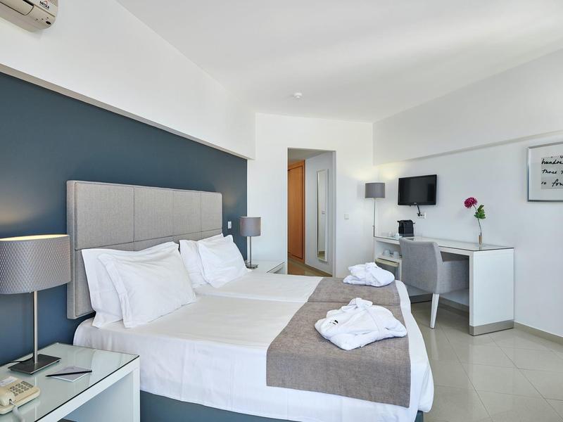 Hell eingerichtetes Schlafzimmer mit Doppelbett, grauem Akzentwand, Schreibtisch und moderner Beleuchtung.