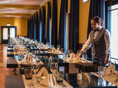 Ein Kellner faltet Servietten in einem elegant gedeckten, langen Restaurant mit dunklen Vorhängen.