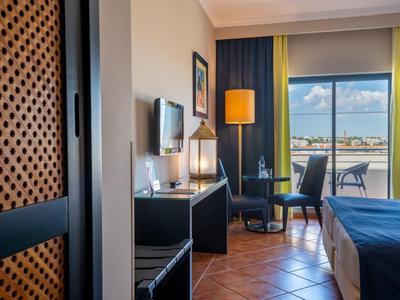 Hell eingerichtetes Hotelzimmer mit Doppelbett, Schreibtisch, Stuhl, Fernseher und Balkon mit Blick aufs Wasser.