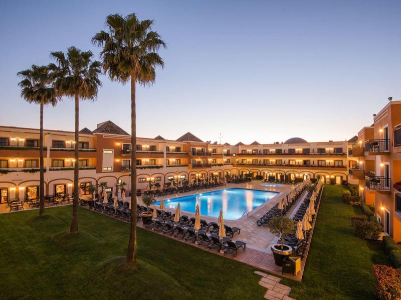 Großes Hotel mit beleuchtetem Pool, Liegestühlen und gepflegtem Grasbereich bei Sonnenuntergang.