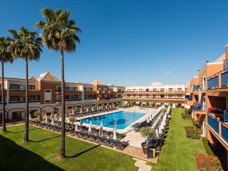 Hotel mit rechteckigem Pool, Liegestühlen, Palmen und klarem blauem Himmel.
