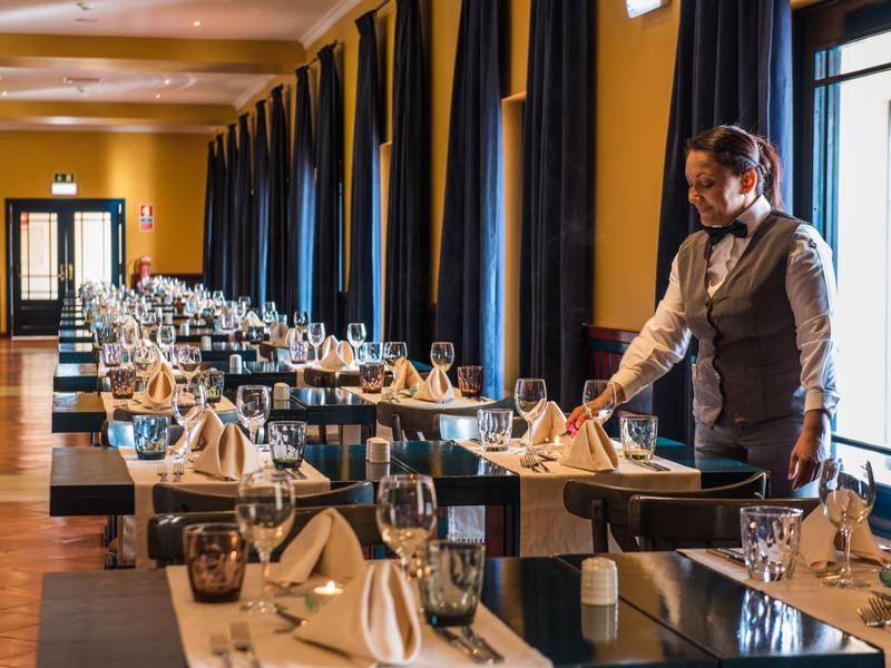 Ein Kellner faltet Servietten in einem elegant gedeckten, langen Restaurant mit dunklen Vorhängen.