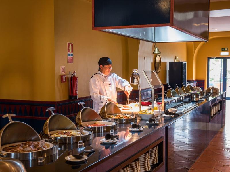 Koch im weißen Mantel bereitet Buffet mit warmen Speisen in hellem Restaurant vor.