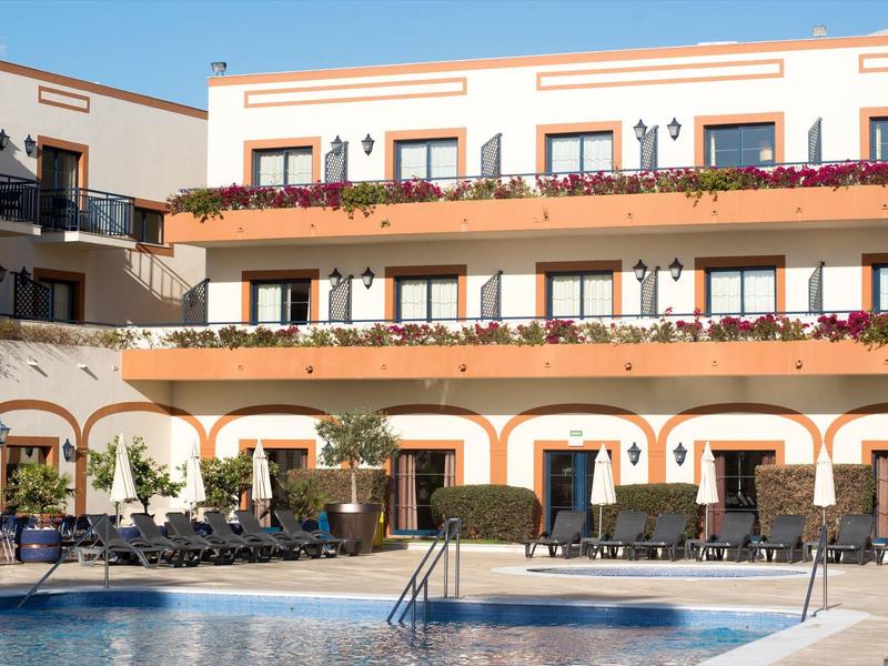 Hotel mit Balkonblumen, Sitzgelegenheiten und Swimmingpool unter klarem blauem Himmel.