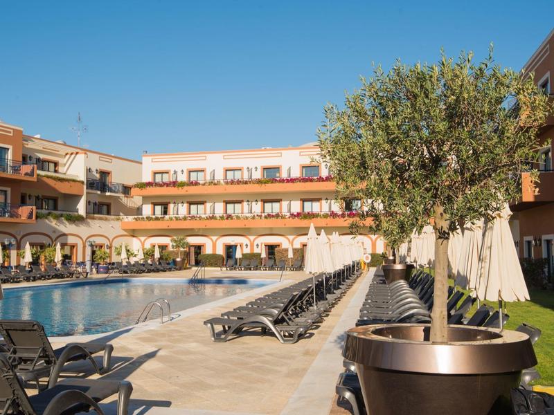 Großer Hotelpool mit Sonnenliegen, Pflanzen und mehrstöckigem Gebäude bei klarem Himmel.