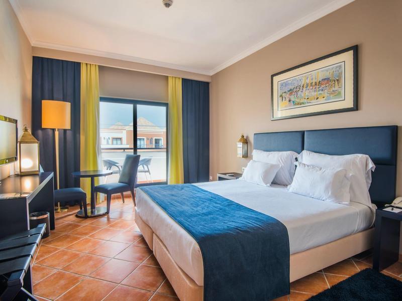 Helles Hotelzimmer mit großem Bett, blauen Akzenten, Schreibtisch und Fenster mit Stadtblick.