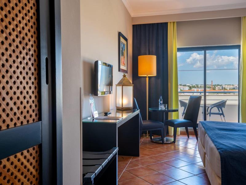 Hell eingerichtetes Hotelzimmer mit Doppelbett, Schreibtisch, Stuhl, Fernseher und Balkon mit Blick aufs Wasser.