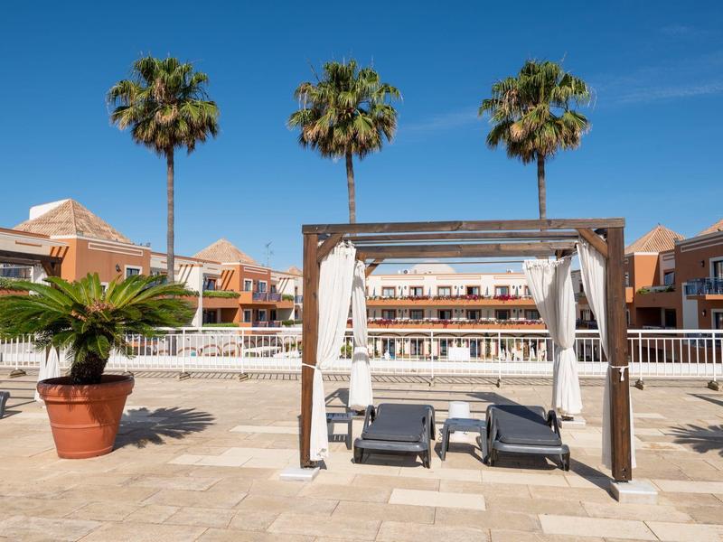Sonnenterrasse mit zwei Liegen unter Holzpergola, drei Palmen und Hotelgebäuden im Hintergrund.
