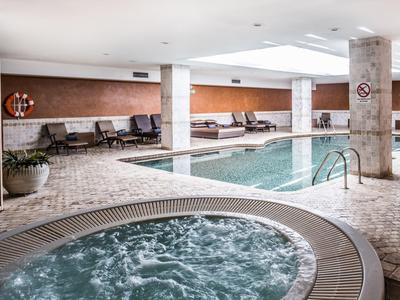 Innenbereich mit Schwimmbecken und rundem Whirlpool, helle Fliesen und Sitzbereiche an der Wand.