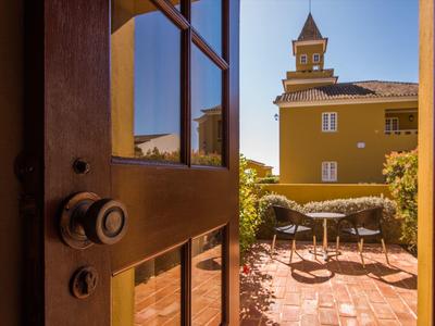 Geöffnete Holztür führt zu sonnigem, gepflastertem Garten mit Tisch, Stühlen und gelbem Gebäude.