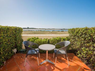 Terrasse mit zwei Stühlen und kleinem Tisch, begrenzt von grünen Hecken, Blick auf Sand und Meer.