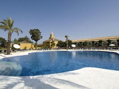 Großer blauer Pool mit Sonnenschirmen, Palmen und gelben Gebäuden unter klarem Himmel.