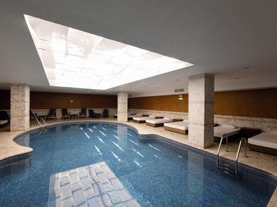 Innenraum Schwimmbad mit blauem Wasser, weißen Säulen, Liegen und großem Oberlicht.