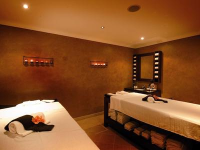 Dunkler Wellnessraum mit zwei Massageliegen, Kerzen und einem beleuchteten Spiegel an der Wand.