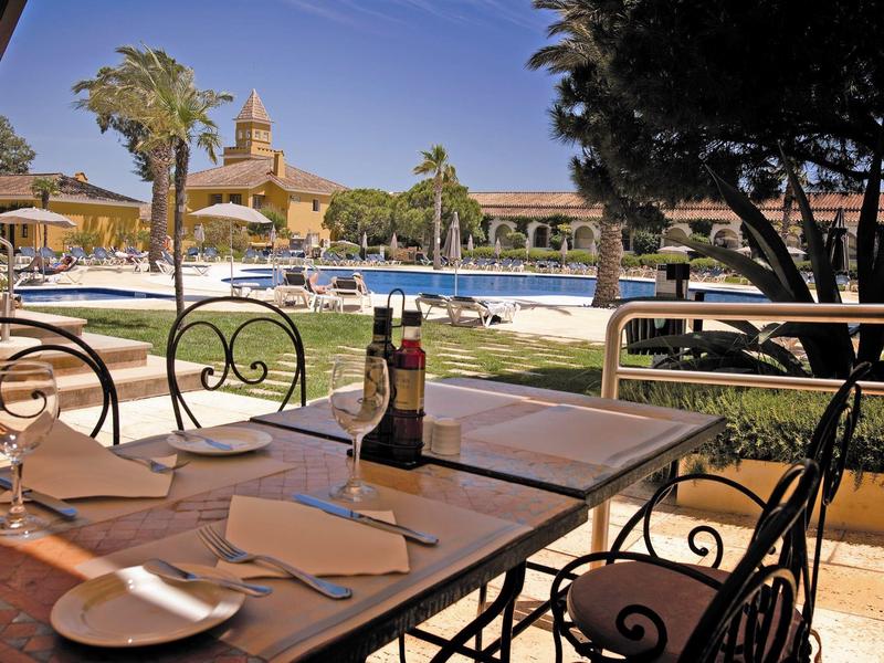 Terrassenrestaurant mit Tisch, Besteck, Weinflasche, Pool und Palmen im Hintergrund an sonnigem Tag.