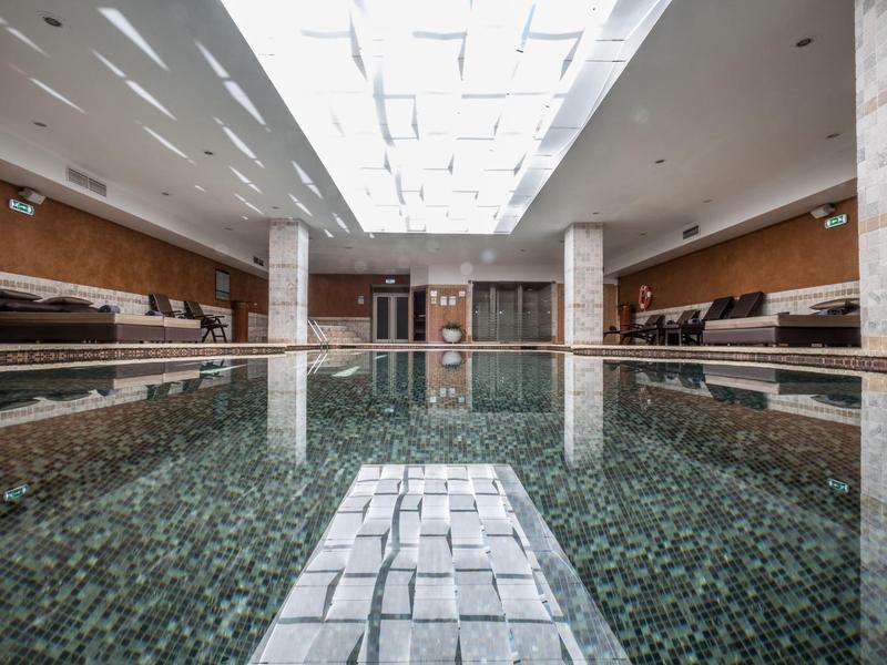 Großes Hallenbad mit ruhigem Wasser, weißen Wänden und Deckenfenstern für Tageslicht.