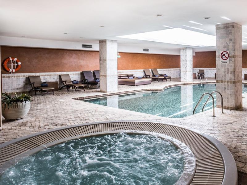 Innenbereich mit Schwimmbecken und rundem Whirlpool, helle Fliesen und Sitzbereiche an der Wand.