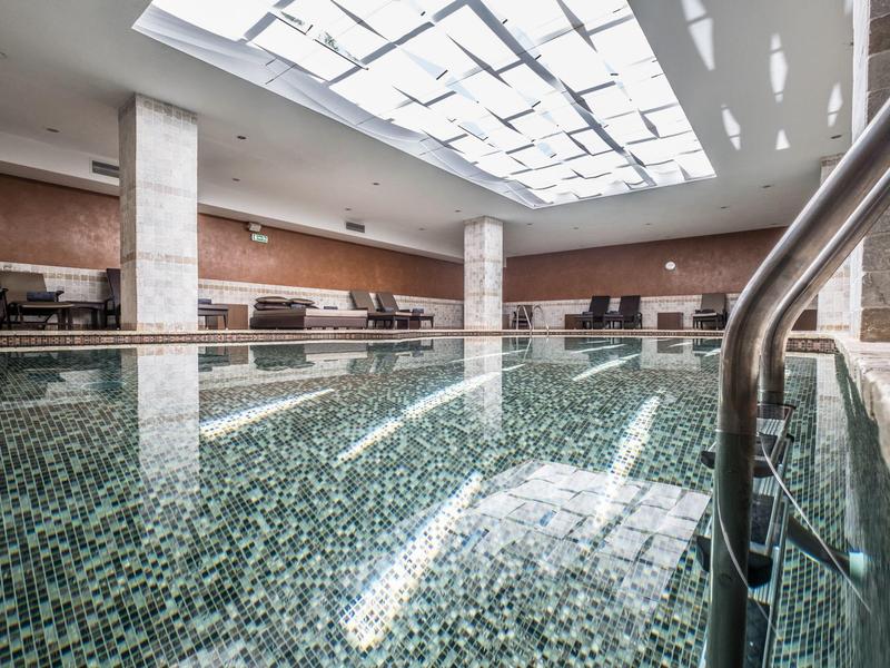 Hell beleuchtetes Hallenbad mit Mosaikfliesen, Edelstahlleiter und großen Fenstern in Decke.