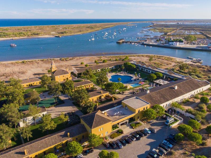 Luftaufnahme einer Küstenanlage mit Bootshafen, Schwimmbad und umgeben von Bäumen und Parkplätzen.