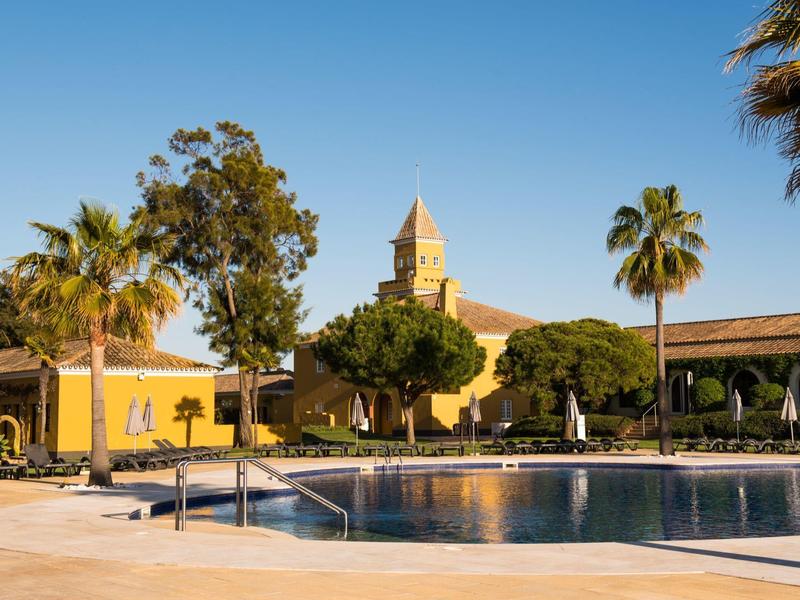 Schwimmbecken mit blauem Wasser, umgeben von gelben Gebäuden, Palmen und klarem Himmel im Hintergrund.