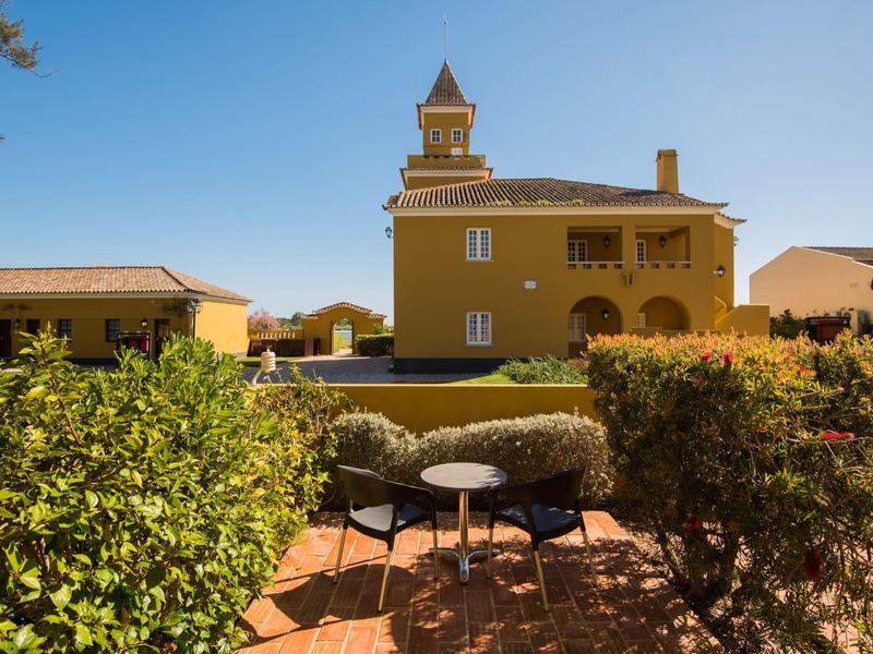 Zwei Stühle mit kleinem Tisch stehen auf Terrasse vor gelbem Haus mit blauen Himmel im Hintergrund.
