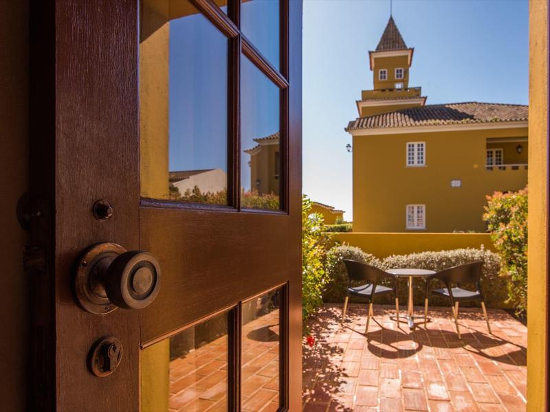 Geöffnete Holztür führt zu sonnigem, gepflastertem Garten mit Tisch, Stühlen und gelbem Gebäude.