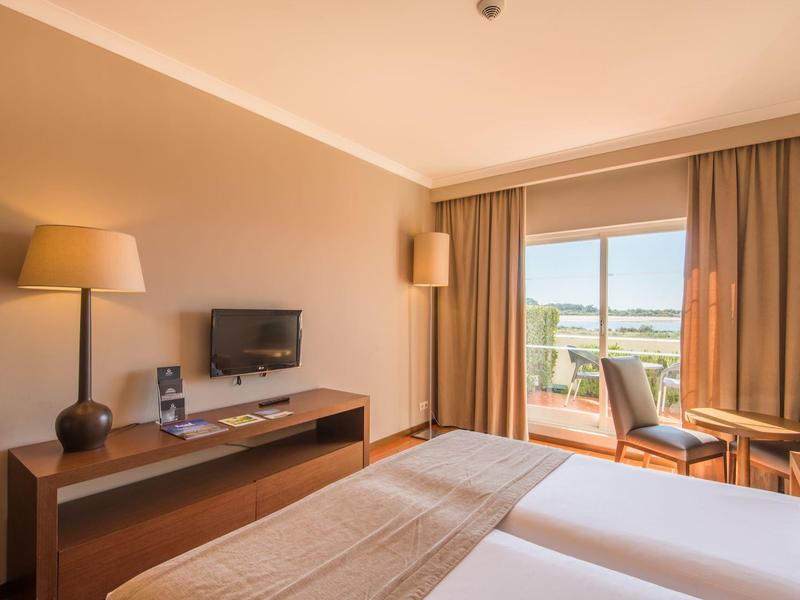 Hell eingerichtetes Hotelzimmer mit zwei Betten, Fernseher, Lampe, Tisch und Blick auf Balkon und Landschaft.