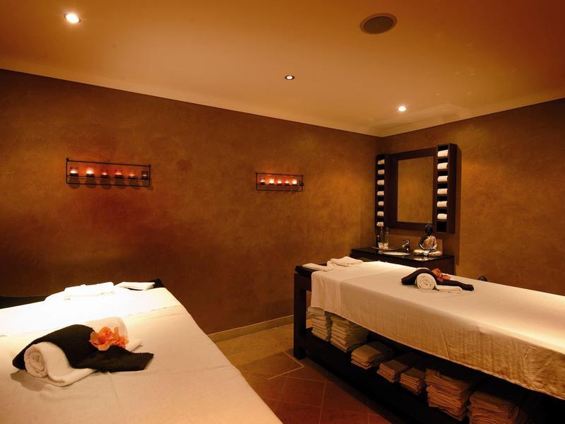 Dunkler Wellnessraum mit zwei Massageliegen, Kerzen und einem beleuchteten Spiegel an der Wand.