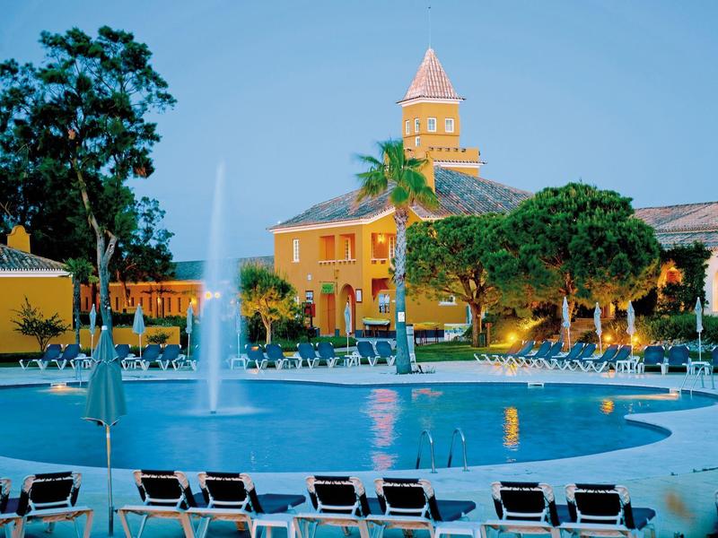 Pool mit Springbrunnen vor einem gelben Gebäude und vielen Liegestühlen im Freien.