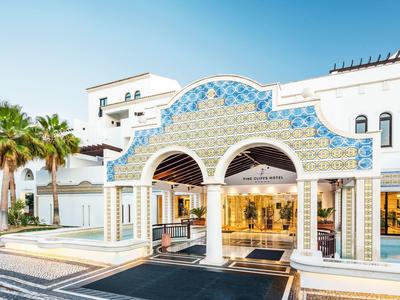 Eingang eines weißen Hotels mit blauen Details und Palmen unter klarem Himmel.