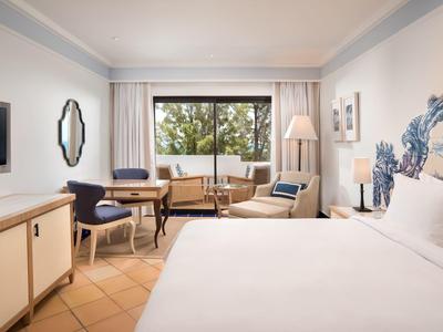 Helles Hotelzimmer mit großem Bett, Schreibtisch, Stühlen und Blick auf grüne Bäume.