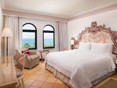 Helles Gästezimmer mit Boxspringbett, Vintage-Kopfteil, zwei Stühlen, Schreibtisch und Meerblick.