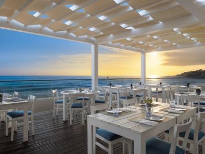 Terras van een strandrestaurant met witte stoelen en tafels, uitzicht op zee bij zonsondergang.
