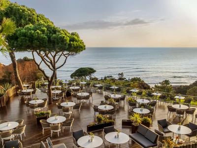 Restaurantterras met ronde tafels en stoelen, omgeven door bomen met uitzicht op zee bij zonsondergang.